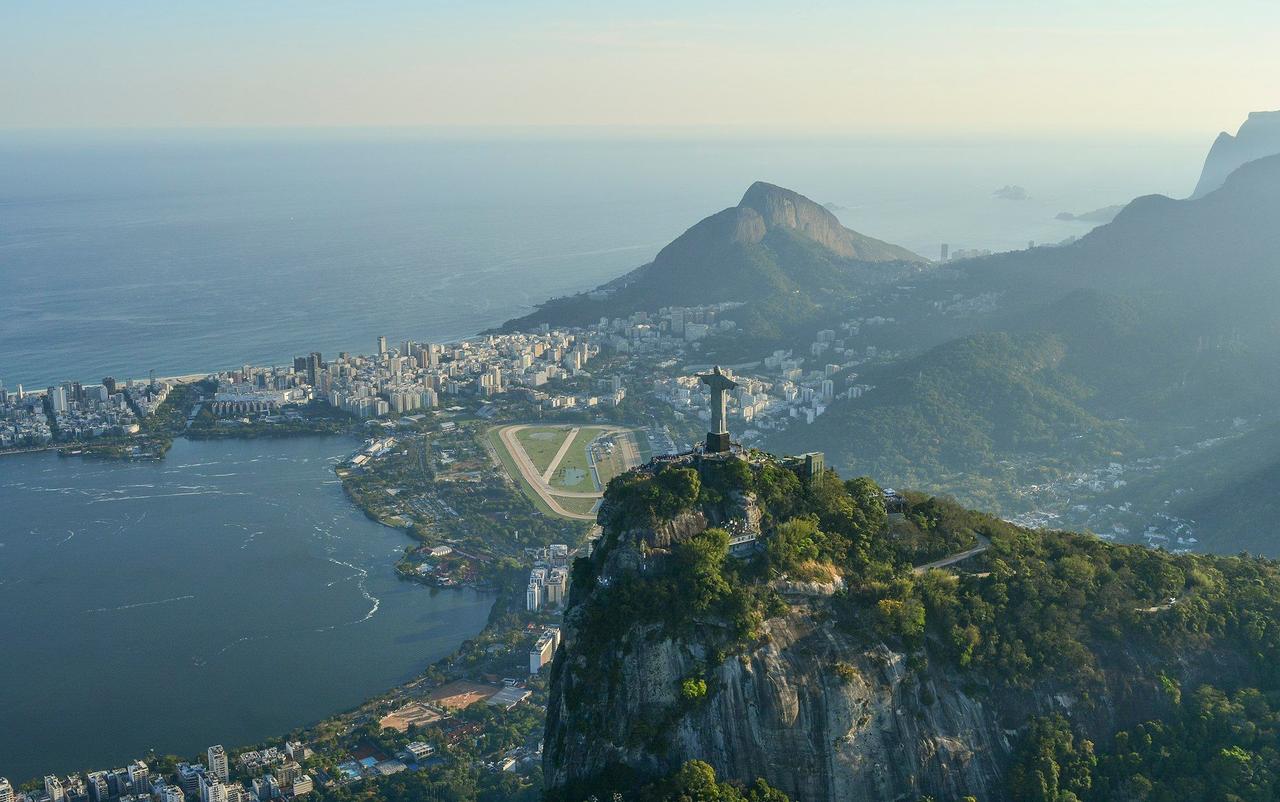 Eine Luftaufnahme einer großen Statue auf einem Berggipfel, mit Blick auf eine Küstenstadt, eine große Lagune und den Ozean.