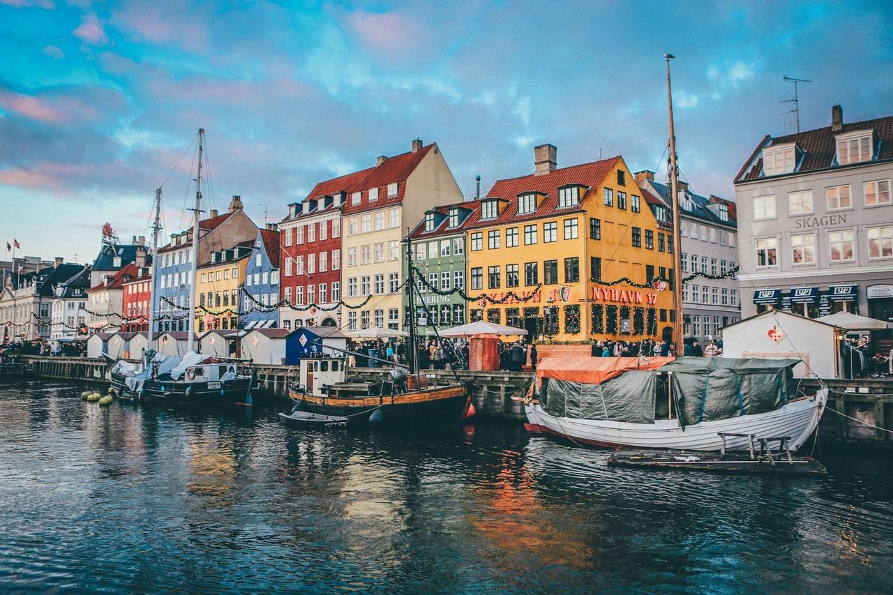 Boote liegen in einem Kanal entlang einer Promenade mit bunten Gebäuden unter einem Pastellhimmel.