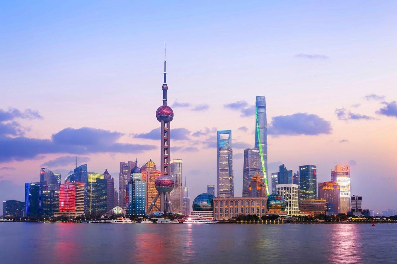 The illuminated skyline of a modern city with futuristic towers reflecting on the water at dusk under a colorful sky.