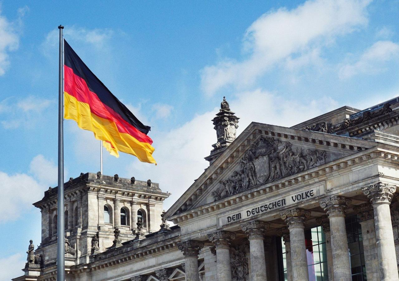 The German flag waves in the wind next to the Reichstag building under a partly cloudy blue sky.