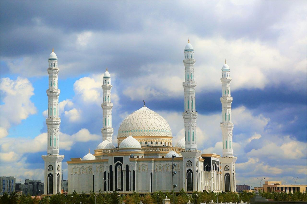 Eine große weiße Moschee mit goldenen und türkisfarbenen Details, einer großen zentralen Kuppel und hohen Minaretten vor einem bewölkten blauen Himmel.