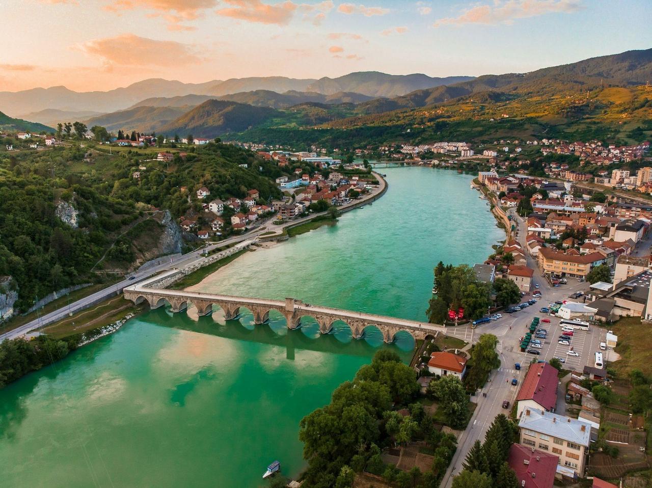 Una vista aérea de un puente de piedra histórico con múltiples arcos cruzando un río turquesa a través de un pueblo rodeado de montañas verdes al atardecer.