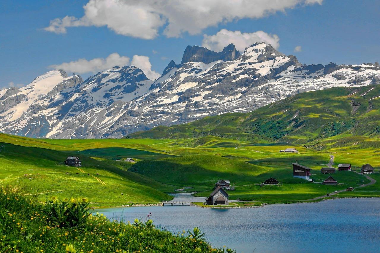 Una vista panorámica de un lago de montaña, colinas verdes ondulantes con pequeñas casas y una gran cordillera nevada al fondo.