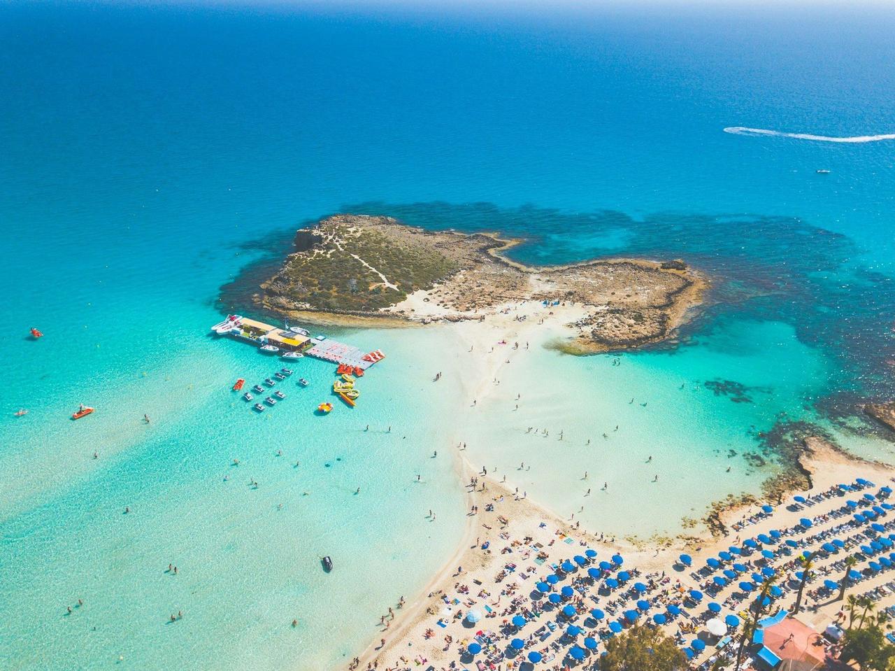 Vista aérea de una playa soleada con gente nadando en aguas cristalinas de color turquesa y relajándose bajo filas de sombrillas azules.