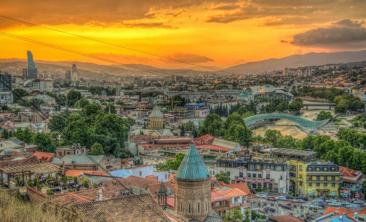 Una vista panorámica de un paisaje urbano con un río, puentes y una mezcla de edificios antiguos y modernos bajo un vibrante atardecer naranja.