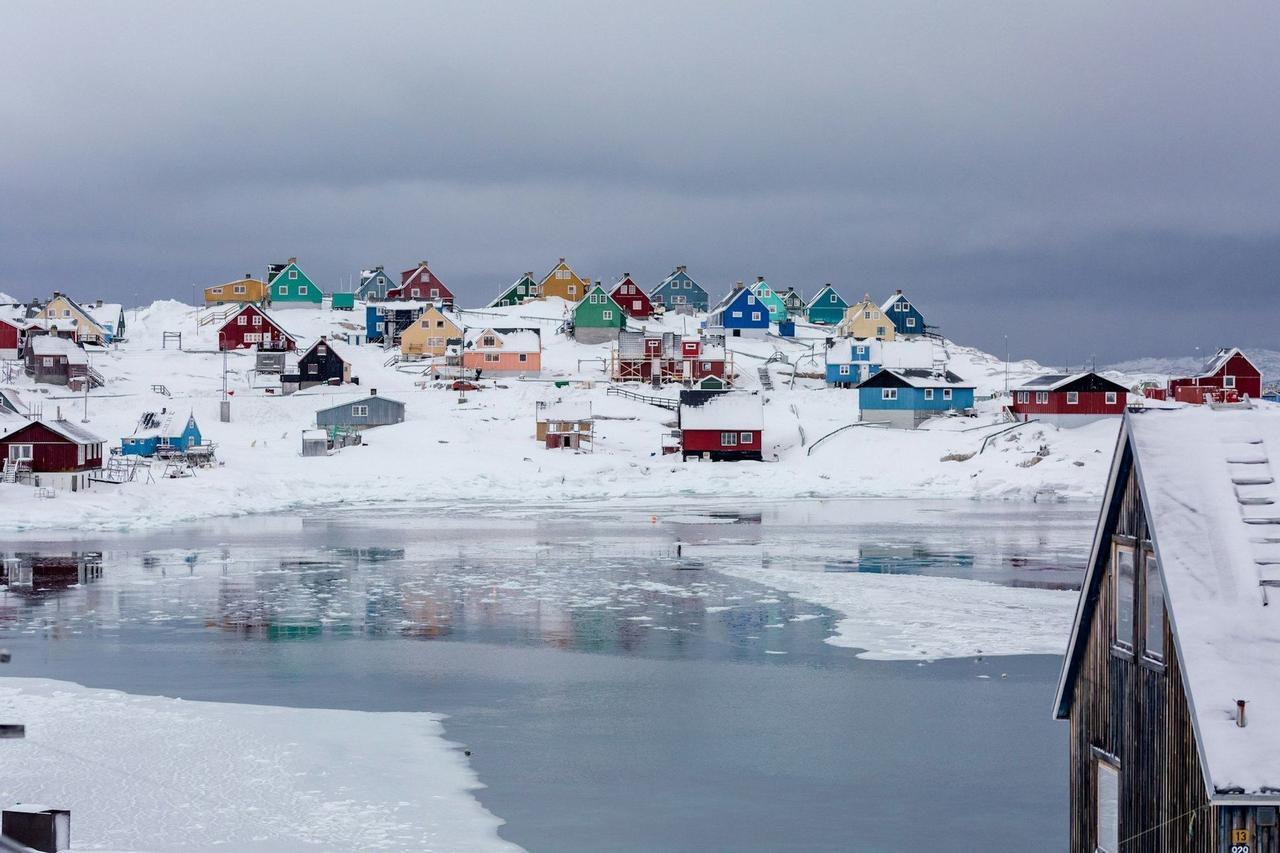 Casas coloridas salpican una ladera nevada junto a una bahía helada bajo un cielo nublado.