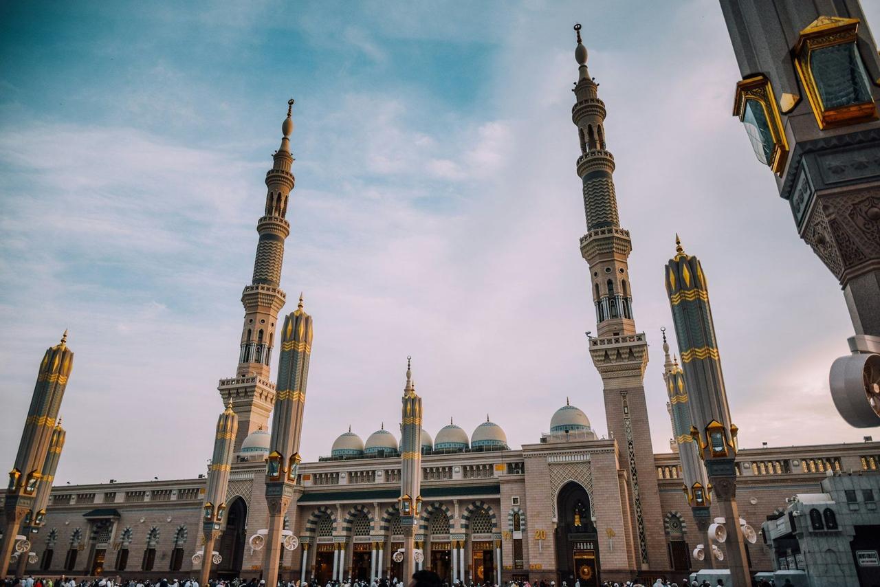Una gran mezquita con altos y ornamentados minaretes y cúpulas, vista desde un ángulo bajo con una multitud reunida enfrente.