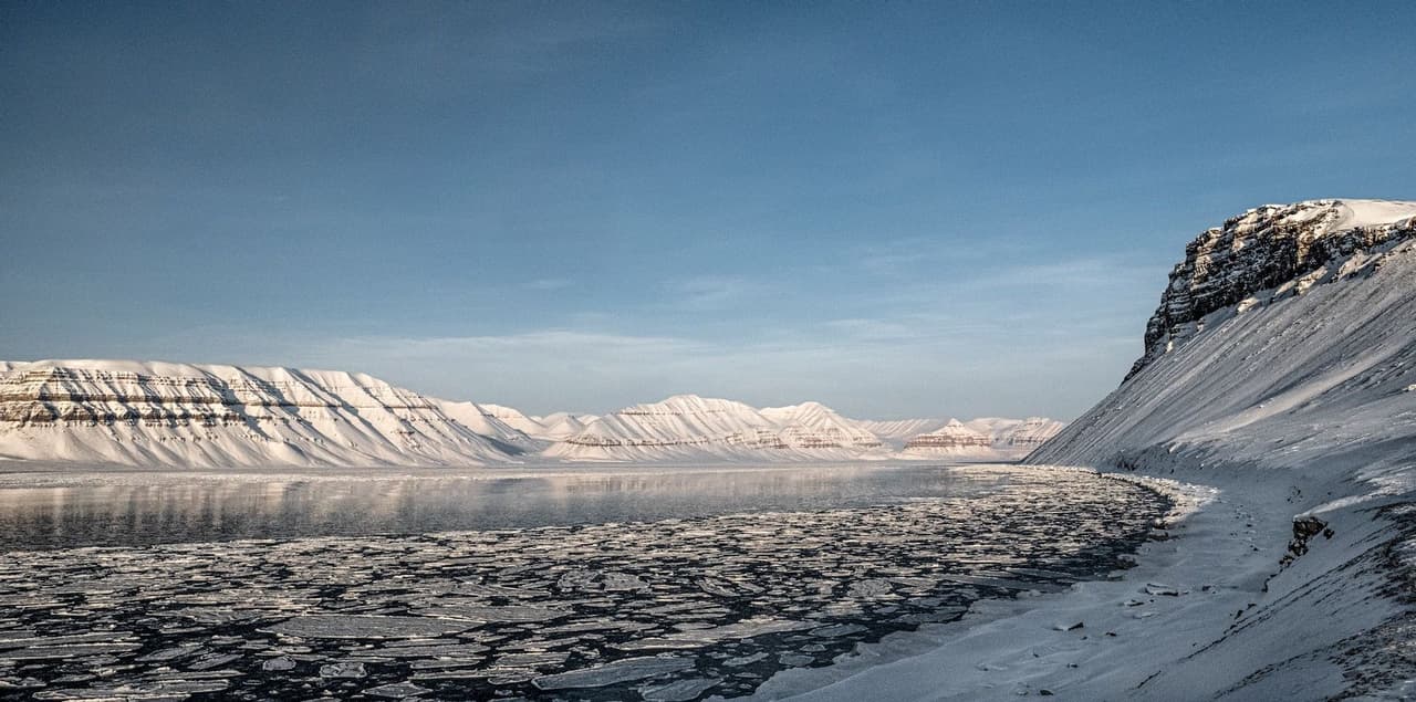 Bloques de hielo flotan en aguas tranquilas en un fiordo rodeado por un vasto paisaje de montañas cubiertas de nieve bajo un cielo azul claro.