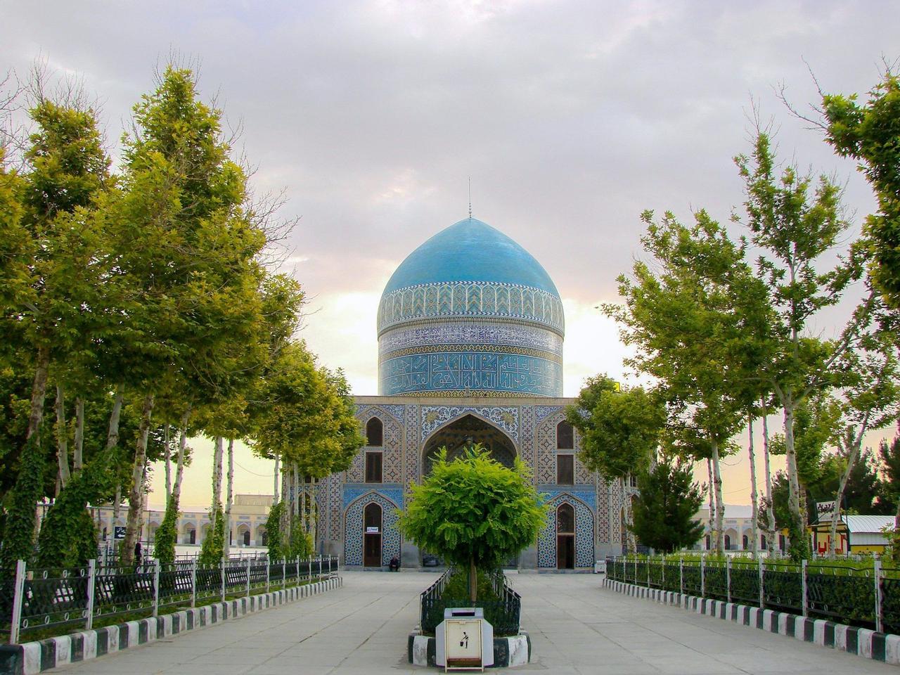 Un sendero de piedra flanqueado por árboles verdes conduce a un edificio ornamentado con una gran cúpula azul claro decorada con intrincados mosaicos.