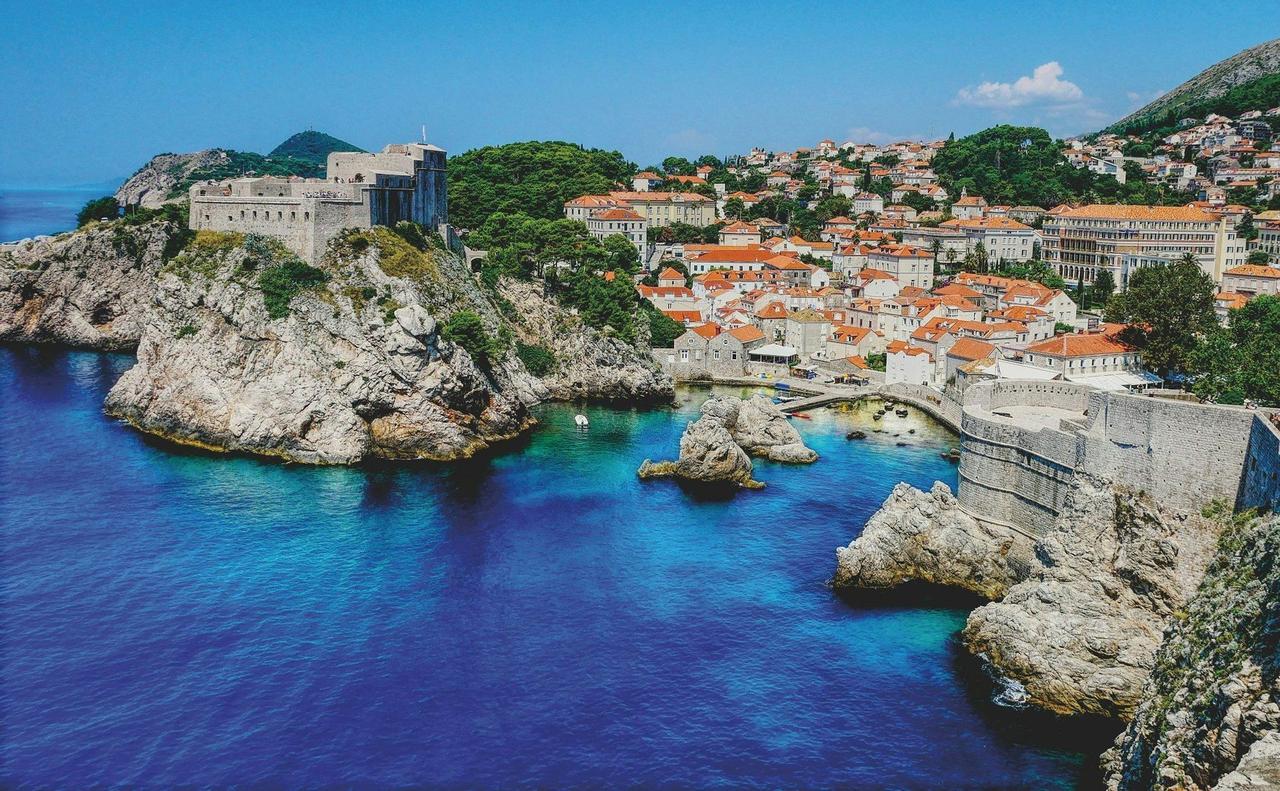 Une ville côtière fortifiée avec des bâtiments aux toits rouges et une forteresse en pierre sur une falaise dominant une baie aux eaux bleues claires.