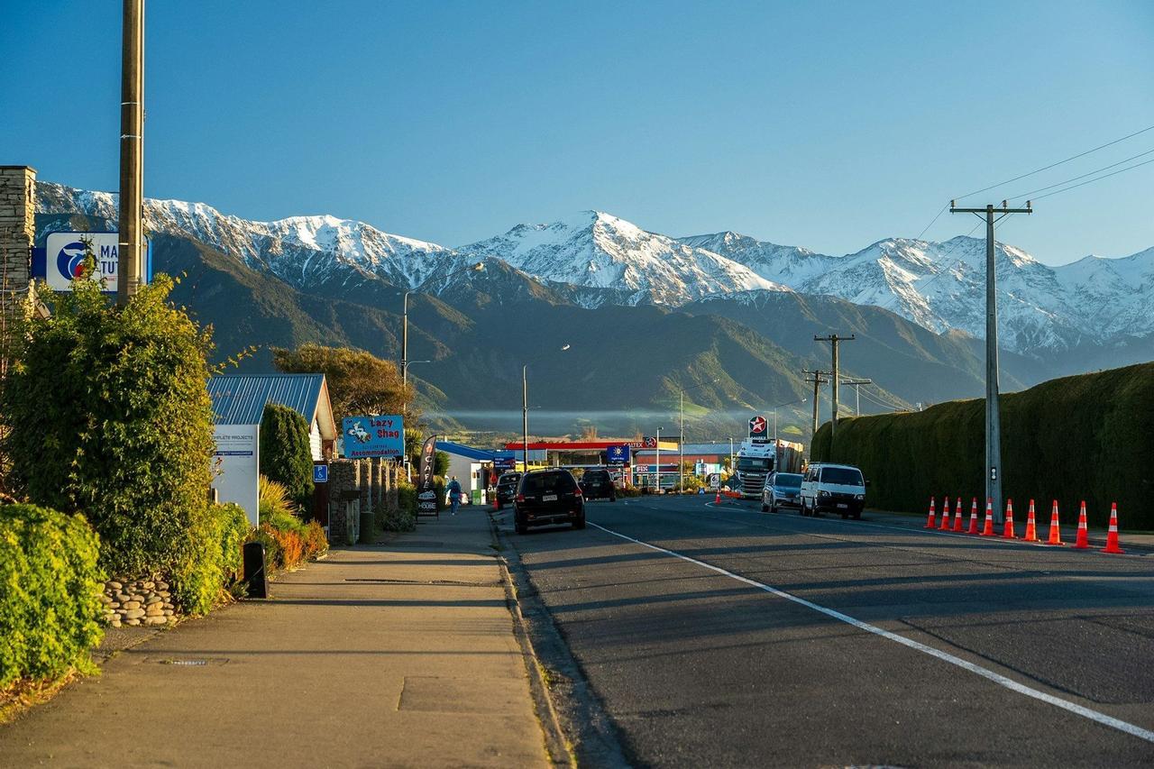Une rue dans une petite ville avec des voitures et une station-service, menant vers une chaîne de montagnes enneigées sous un ciel bleu clair.