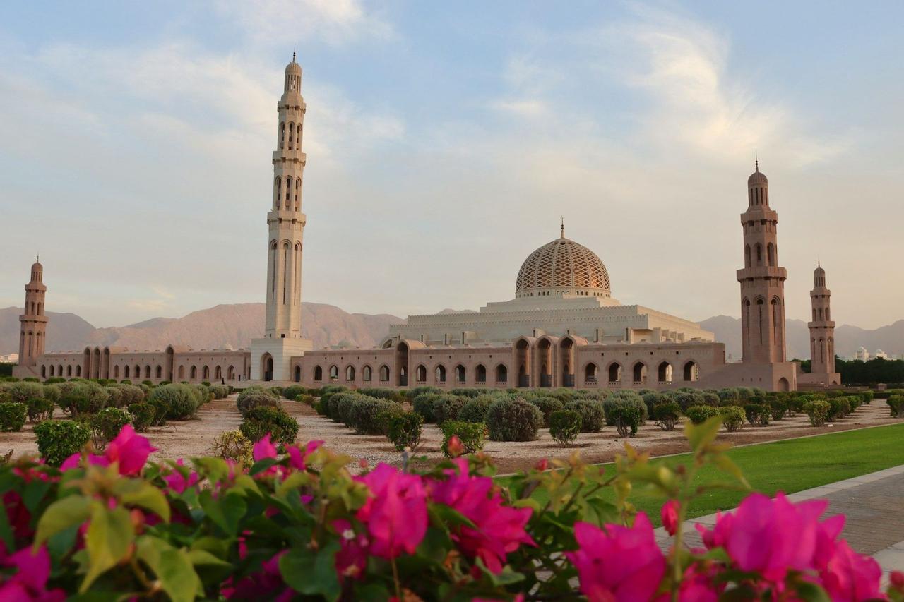 Une grande mosquée avec un dôme et des minarets est visible à travers un jardin avec des fleurs roses au premier plan et des montagnes en arrière-plan.