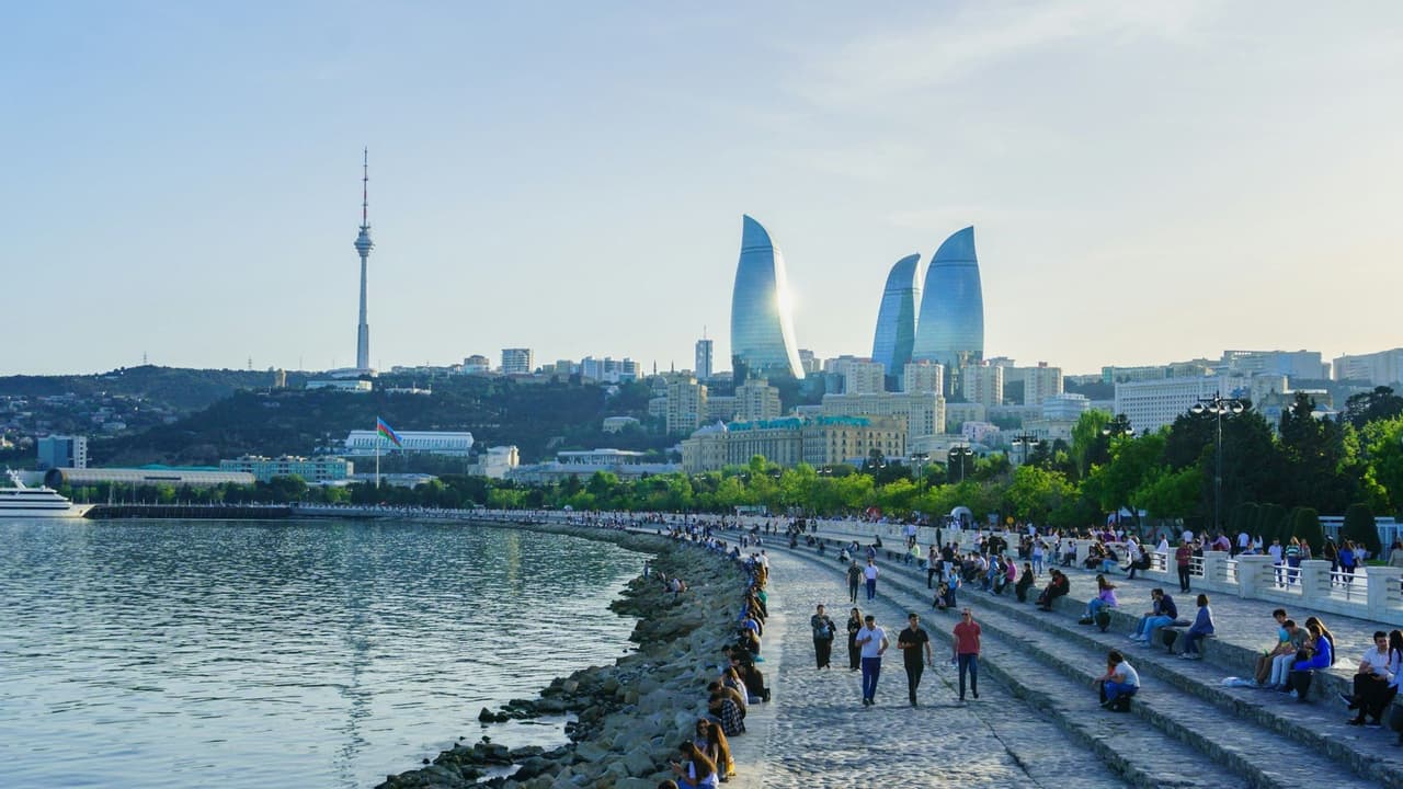 A sunny view of a modern city skyline from a busy waterfront promenade where people are walking and sitting by the water.