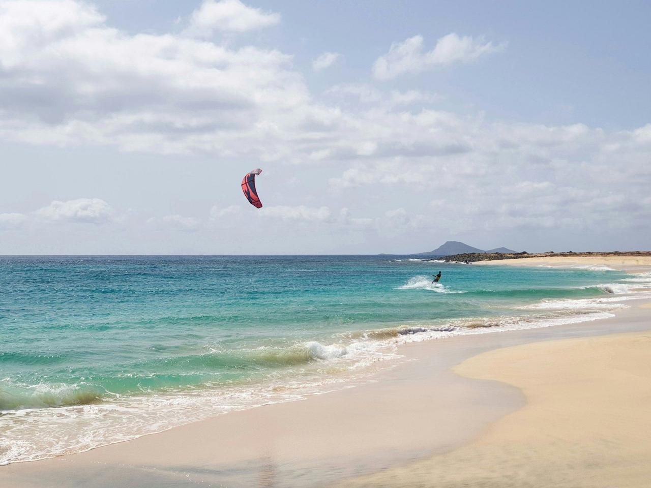 A person kitesurfing in turquoise water near a sandy beach, with a red kite against a partly cloudy sky.