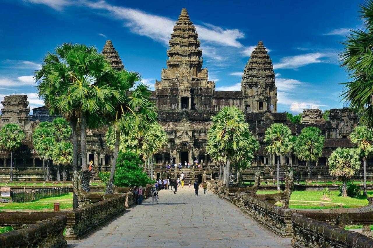 A long stone causeway leads towards a large, ancient temple with multiple towers, flanked by green palm trees under a blue sky.
