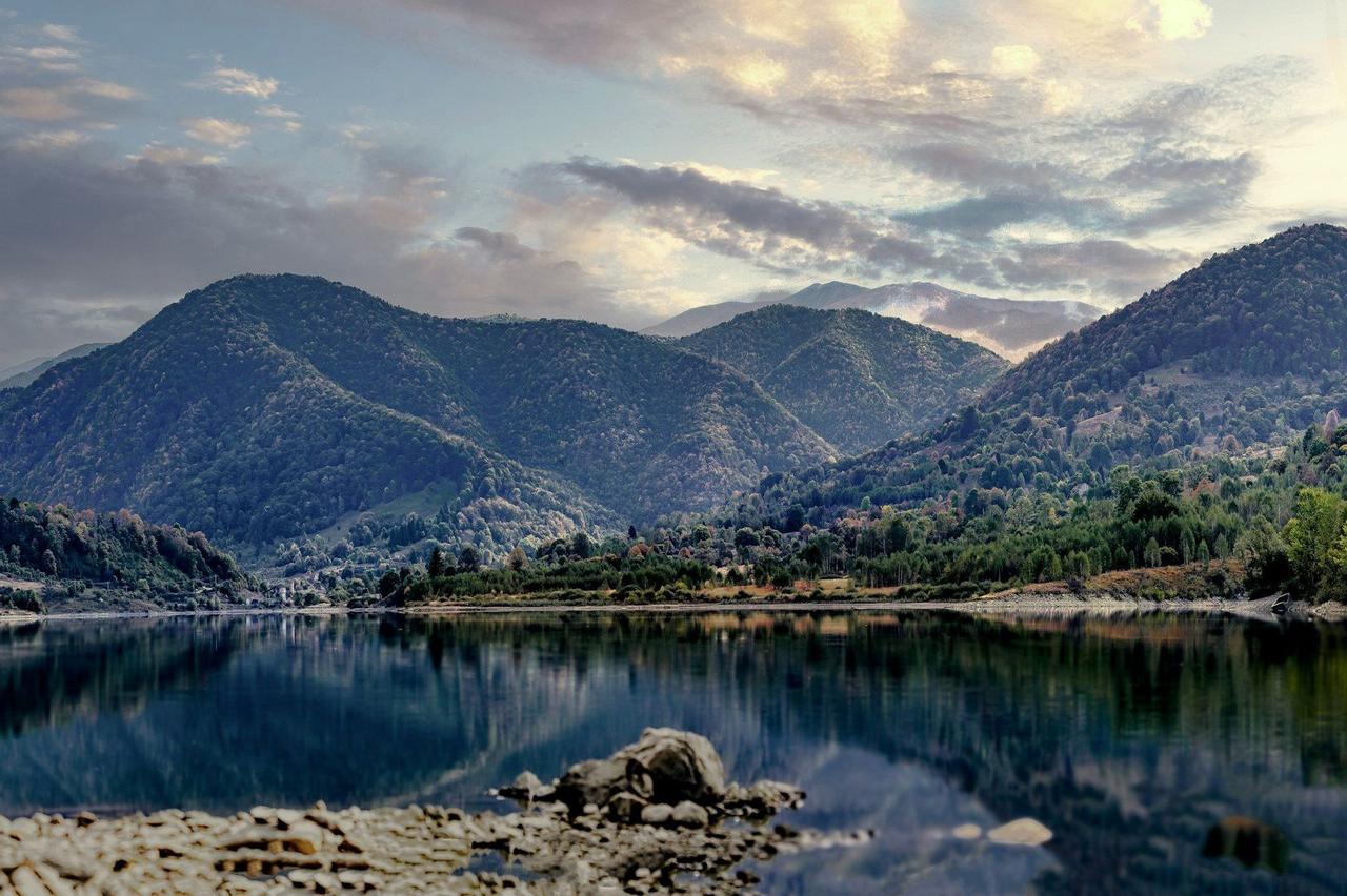 A calm lake reflects tree-covered mountains under a cloudy sky, with a rocky shore visible in the foreground.