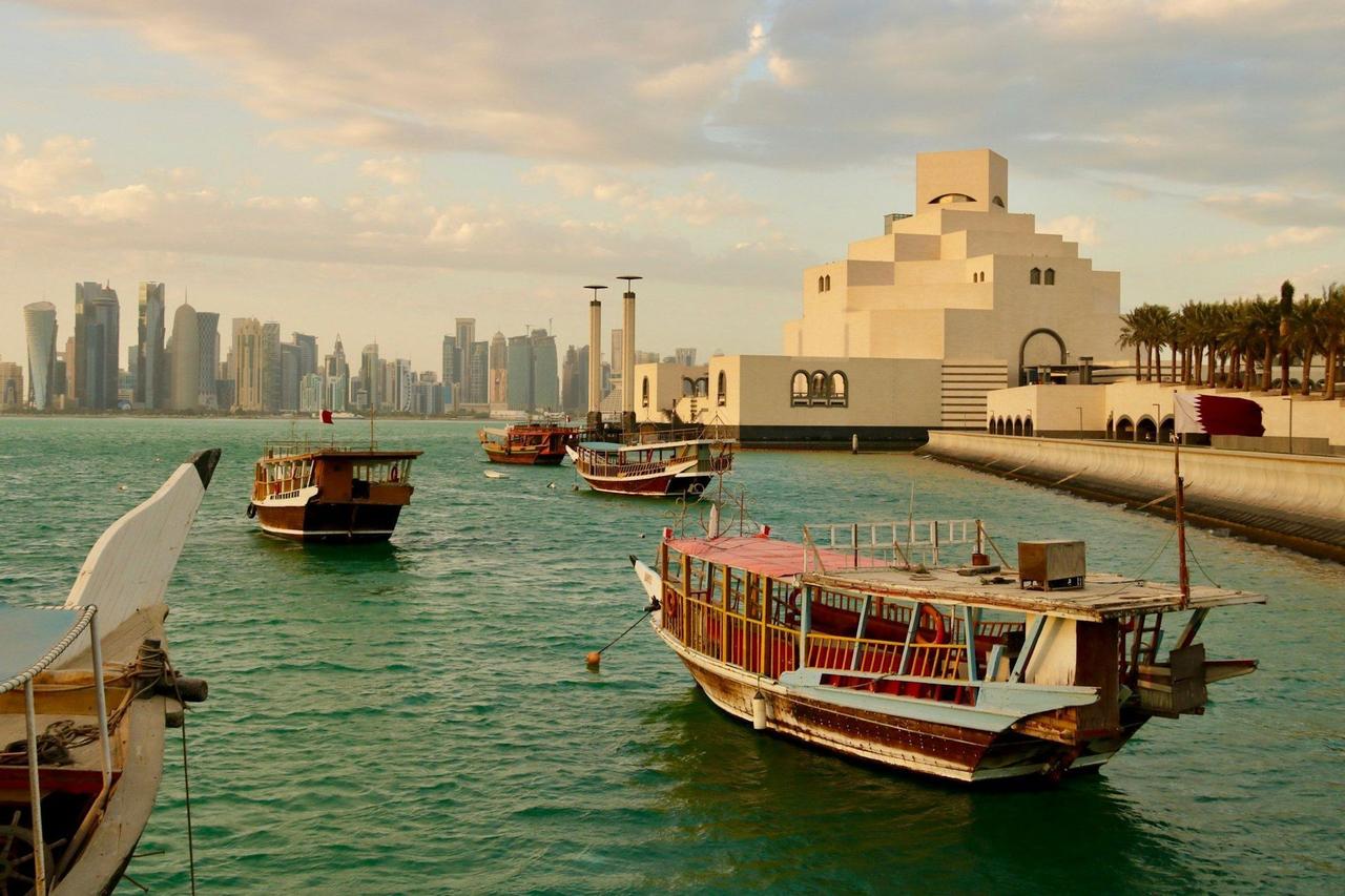 Traditional wooden boats floating in a bay, with a modern city skyline and a large geometric museum in the background.