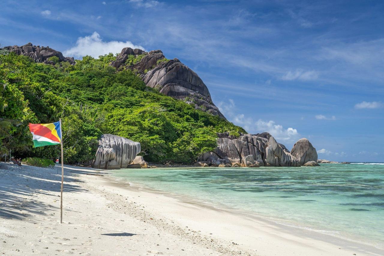 La bandera de Seychelles ondea en una playa de arena blanca junto a aguas turquesas, con una exuberante ladera rocosa y verde de fondo.