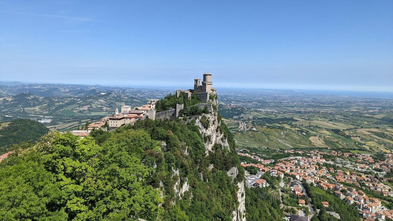 A medieval castle sits atop a tree-covered cliff, overlooking a sprawling valley of towns and rolling green hills under a blue sky.