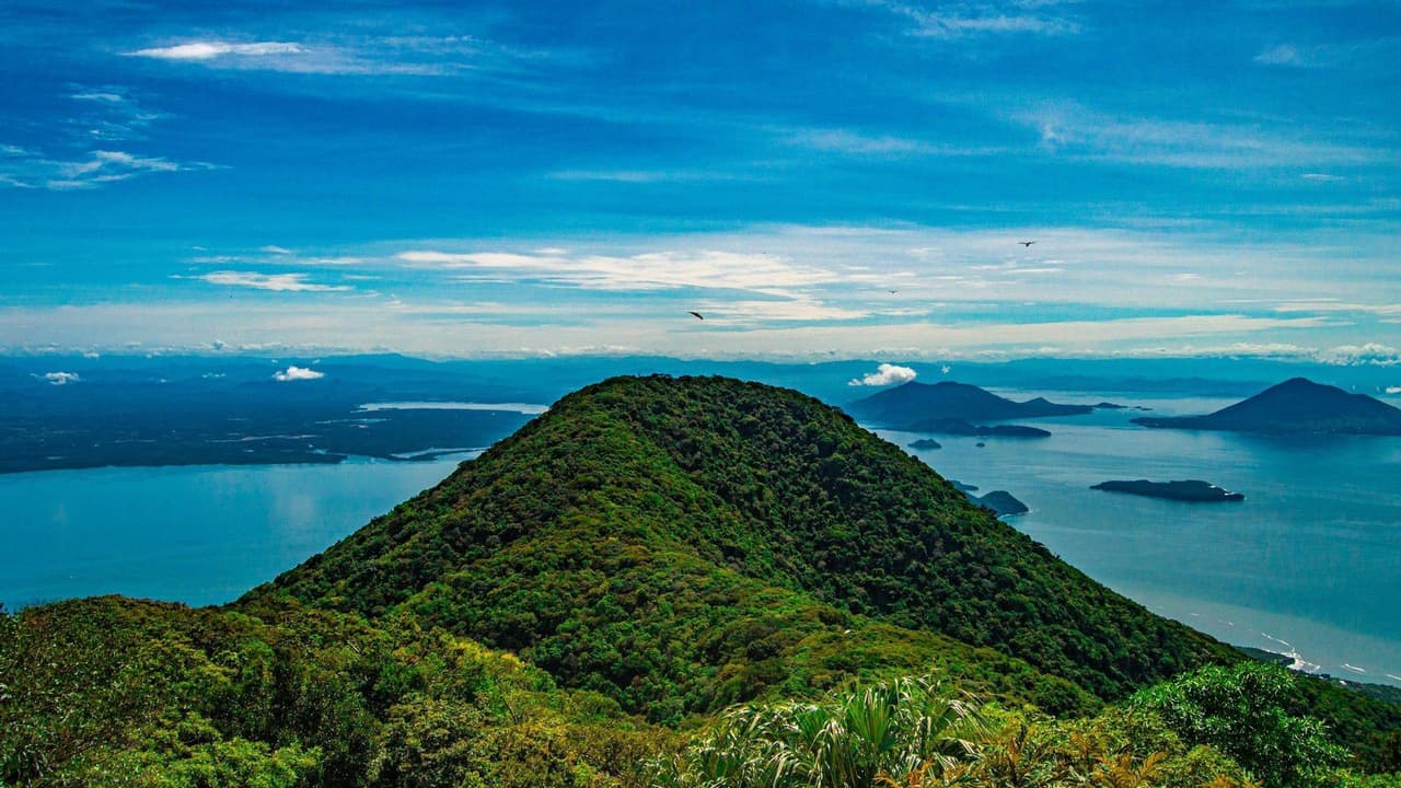 A lush, forest-covered mountain peak rises above a large body of blue water dotted with other islands under a blue sky.