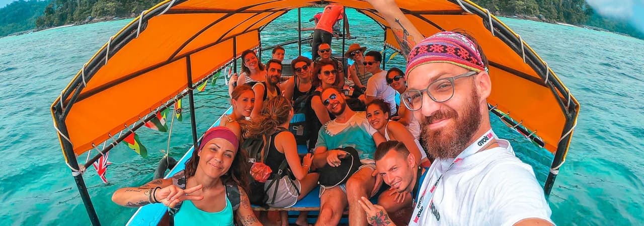 A WeRoad group trip smiles for a selfie on a long-tail boat, sailing on turquoise water near a lush green island.