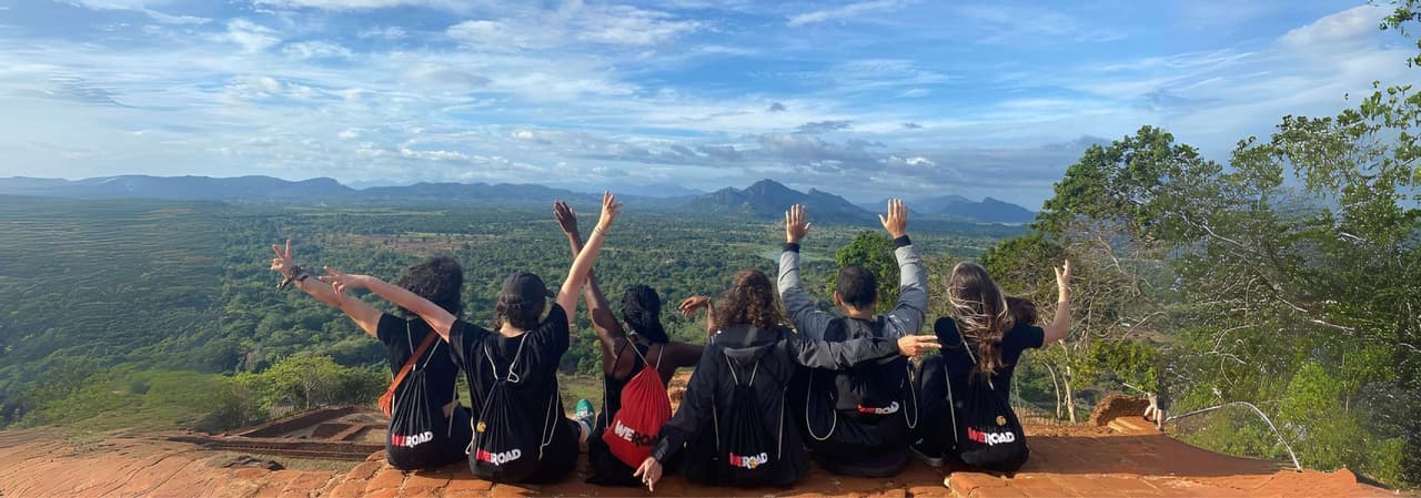 Gruppo WeRoad visto da dietro con le braccia alzate, seduto su un punto panoramico che domina una vasta valle verde e montagne lontane.