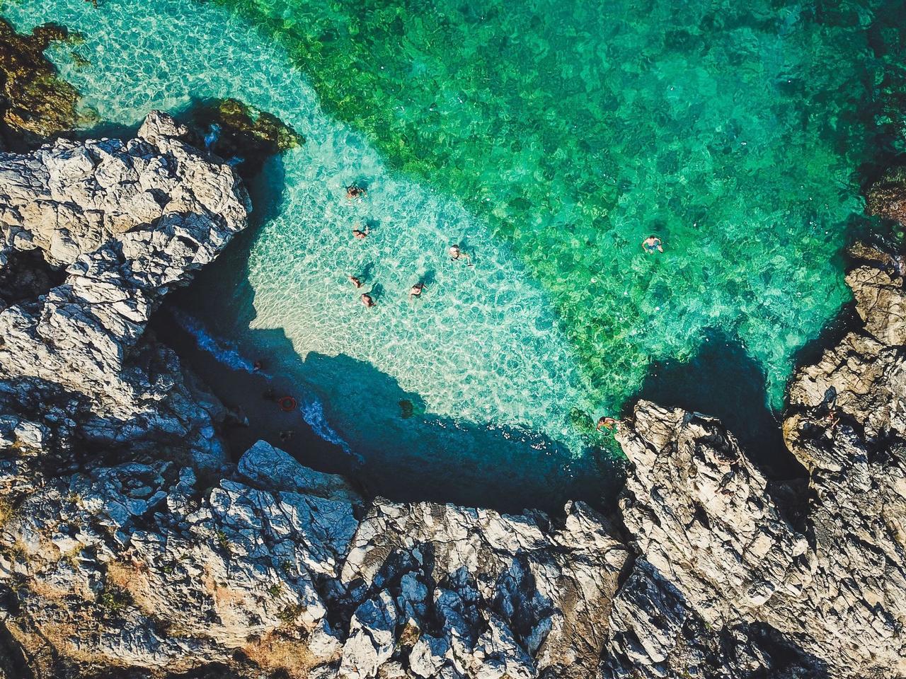 Una vista aérea de personas nadando en las claras aguas turquesas de una cala rocosa.