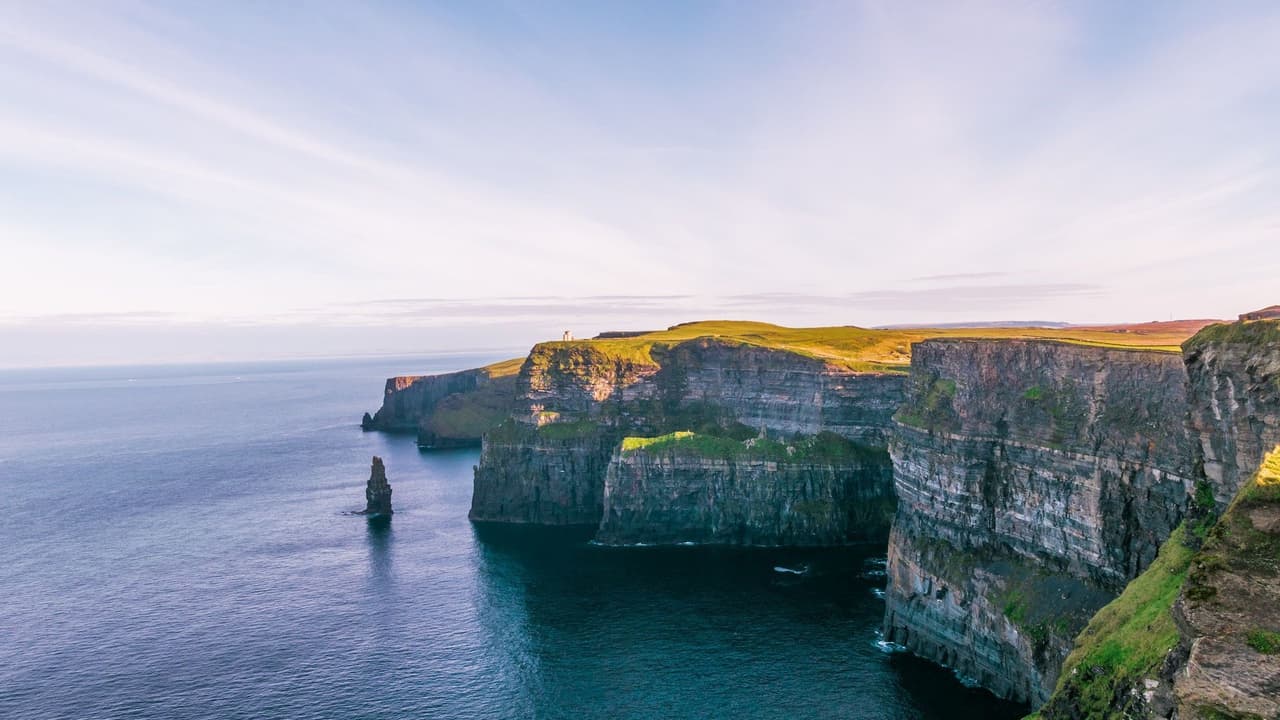 Une vue aérienne de vastes falaises vertes rencontrant l'océan, avec une tour en pierre sur un promontoire et du bétail paissant dans un champ.