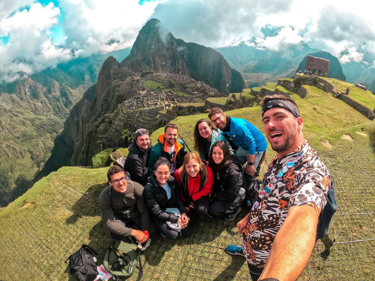 Un grupo de WeRoad se toma una selfie sonriente en un mirador cubierto de hierba con ruinas de montañas antiguas al fondo.