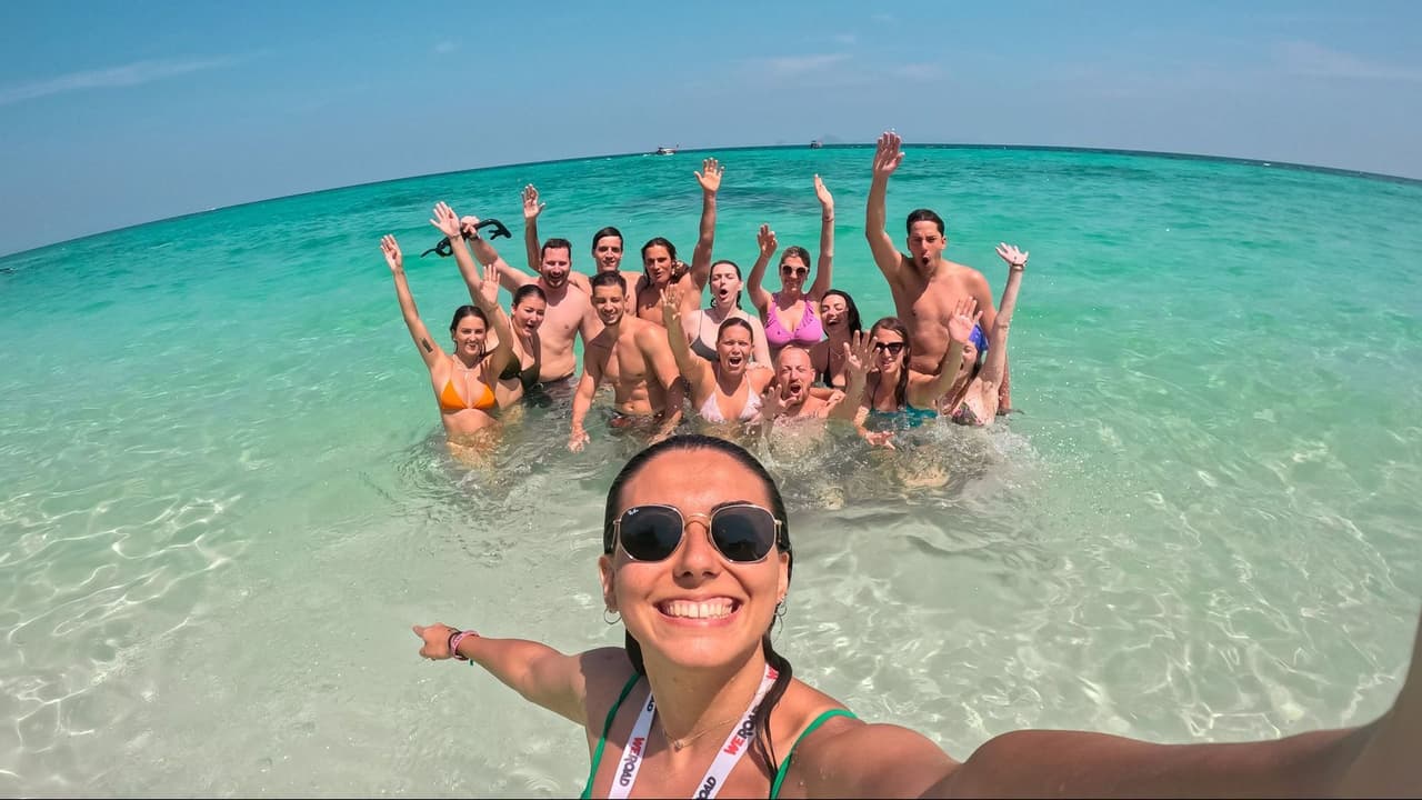 Una mujer se toma una selfie con su grupo de viaje de WeRoad mientras nadan y saludan en aguas cristalinas de color turquesa.