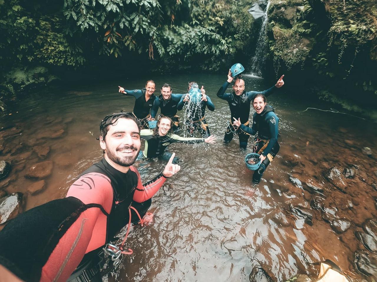 Eine WeRoad-Gruppenreise in Neoprenanzügen macht ein lachendes Selfie, während sie in einem Fluss steht, umgeben von üppigem Grün und einem Wasserfall.