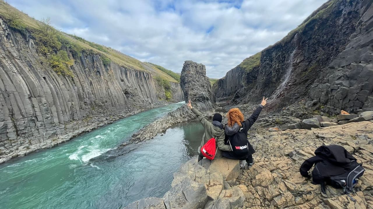 Two people from a WeRoad group trip sit on a rock ledge with arms raised, overlooking a turquoise river in a basalt column canyon.