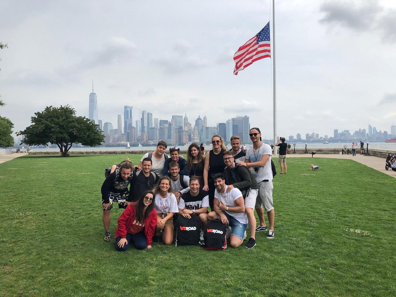A WeRoad group trip poses for a photo on a grassy lawn, with a city skyline and an American flag in the background.