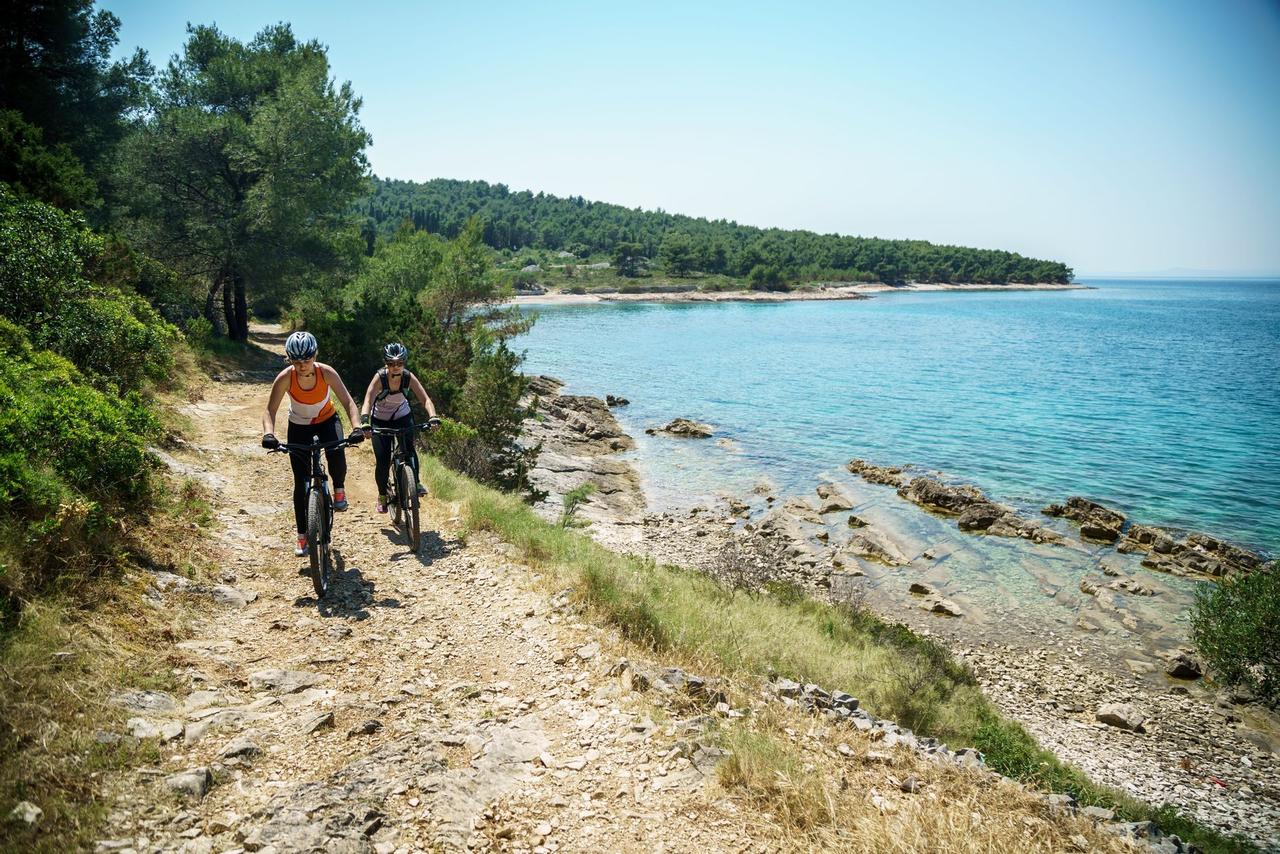 Due donne di un viaggio di gruppo WeRoad pedalano in mountain bike su un sentiero costiero roccioso accanto a un mare turchese sotto un cielo sereno.