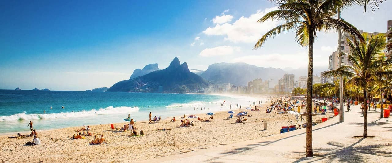 Una giornata di sole su una spiaggia affollata con acqua turchese, uno skyline cittadino e grandi montagne che si stagliano sullo sfondo.