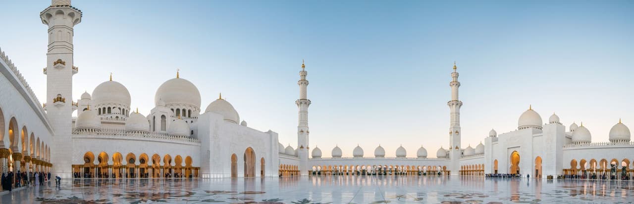 Una grande moschea bianca con cupole e archi si riflette sul pavimento in marmo lucido del cortile sotto un cielo limpido.