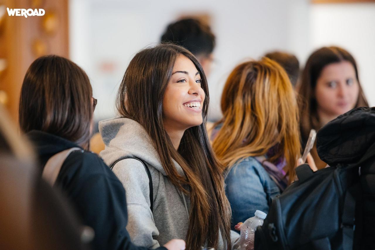 A young woman in a grey hoodie smiles while looking away, surrounded by other people on a WeRoad group trip.