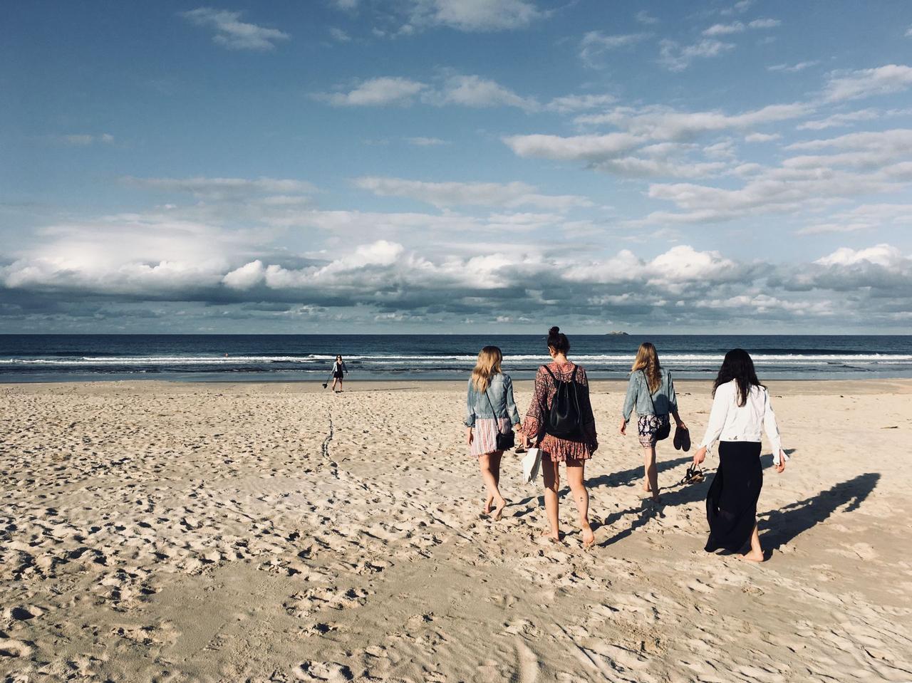 Un voyage de groupe WeRoad de quatre femmes marchant pieds nus sur une plage de sable vers l'océan sous un ciel nuageux.