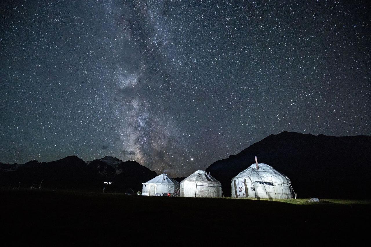 Tre yurte illuminate riposano in un campo ai piedi delle montagne sotto un cielo stellato con la Via Lattea.