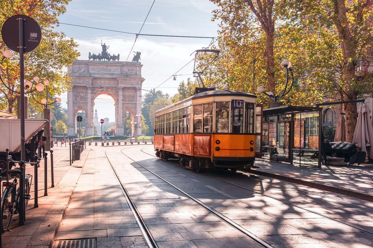 Un tranvía vintage naranja sobre vías pasa junto a un gran arco de triunfo en una calle urbana soleada y arbolada en otoño.