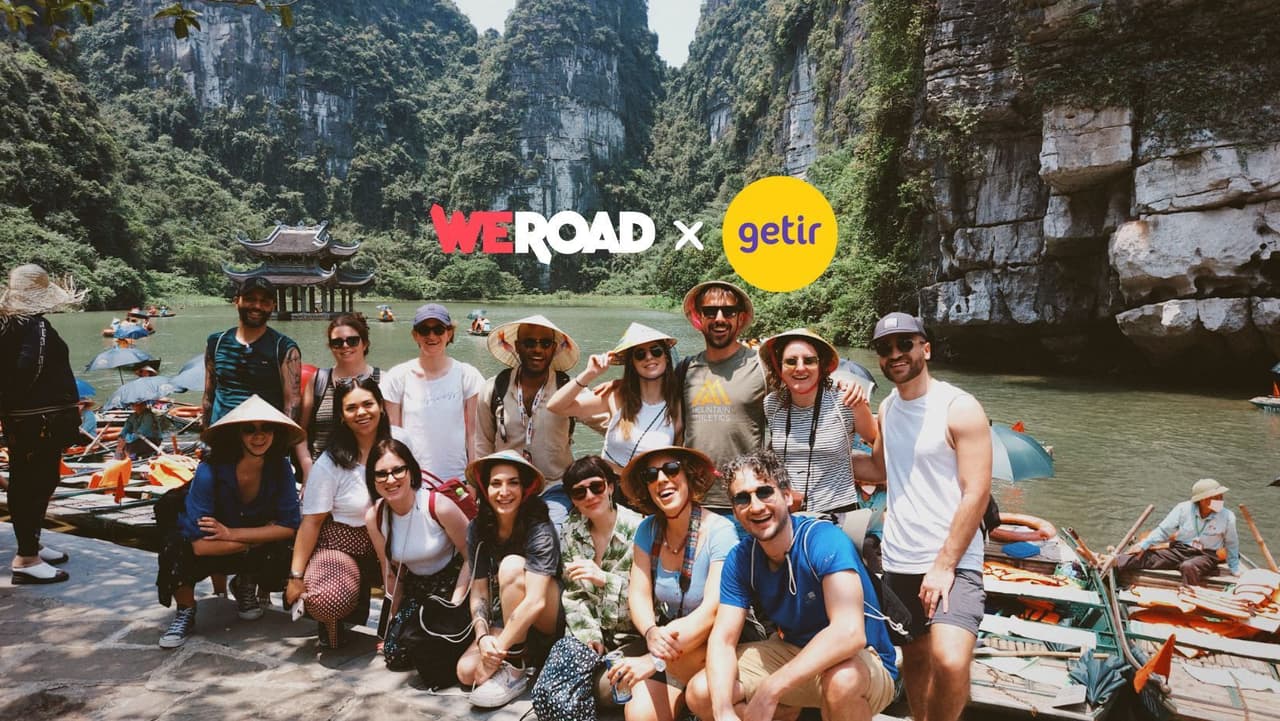 A WeRoad group trip poses for a photo by a river with boats, set against a backdrop of lush mountains and a pagoda.