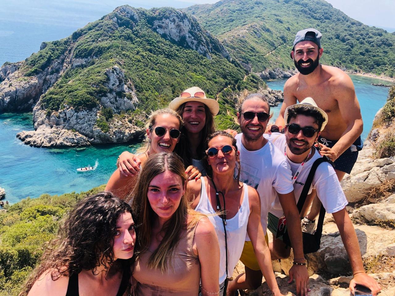 Un viaje en grupo de WeRoad posa para una selfie en un acantilado soleado con vistas a una bahía turquesa y colinas verdes.