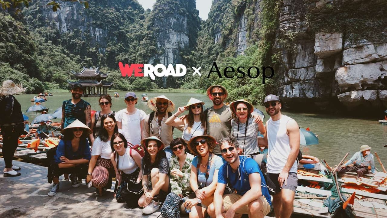 A WeRoad group trip poses for a photo on a riverbank, with lush limestone mountains and traditional boats in the background.