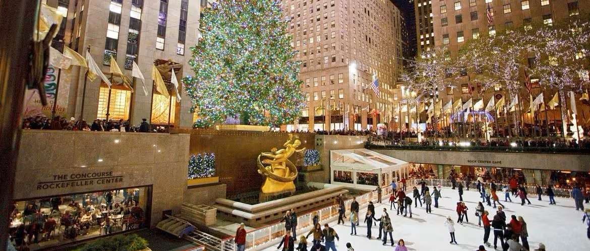 People ice-skating on an outdoor rink at night, with a large lit Christmas tree and tall city buildings in the background.