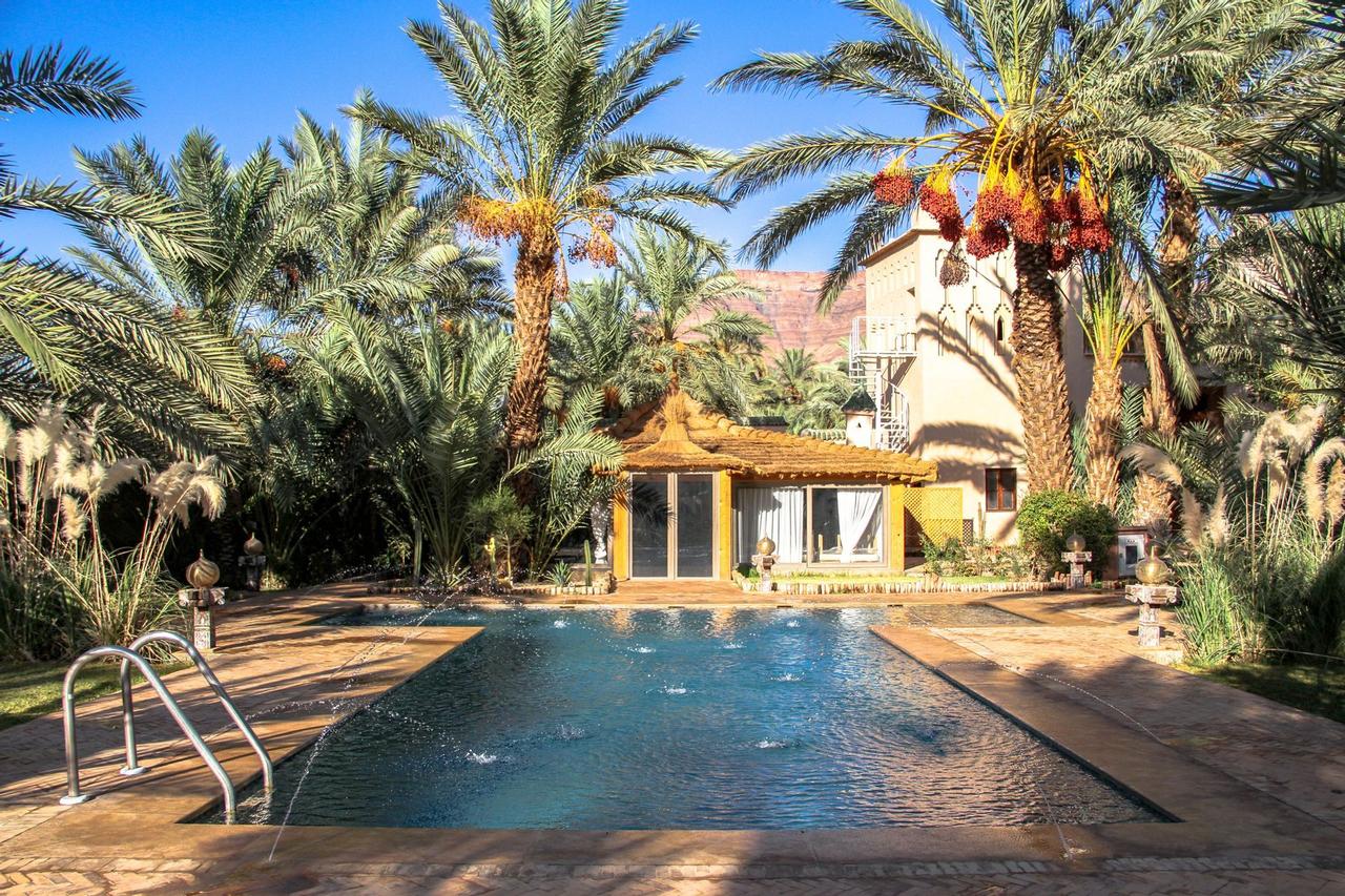 Ein Swimmingpool mit kleinen Springbrunnen liegt in einem sonnigen Innenhof, umgeben von üppigen Palmen und Gebäuden.