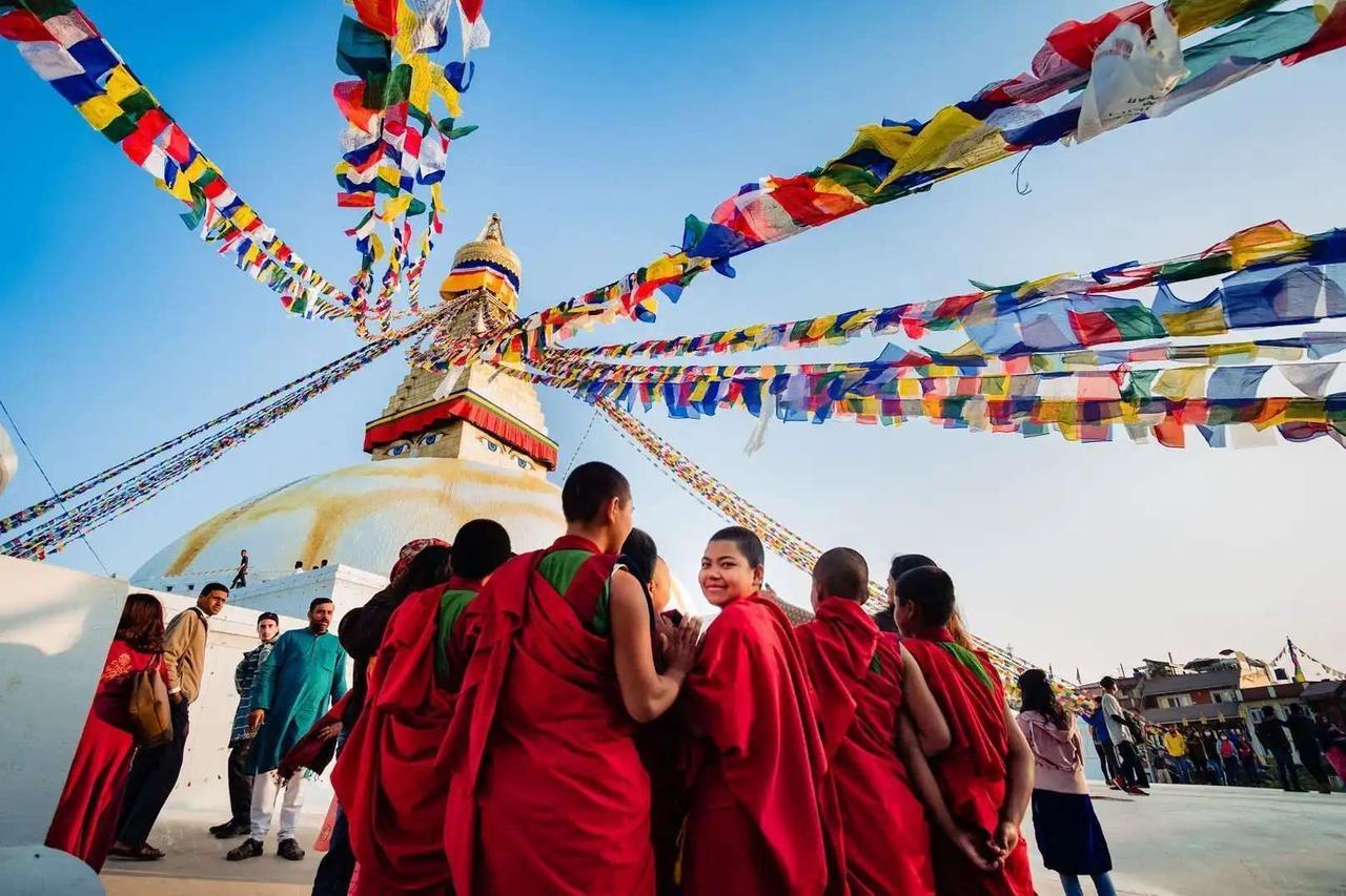 Un groupe de moines bouddhistes vêtus de robes rouges se tient devant un grand stupa blanc orné de drapeaux de prière colorés flottant dans un ciel bleu.