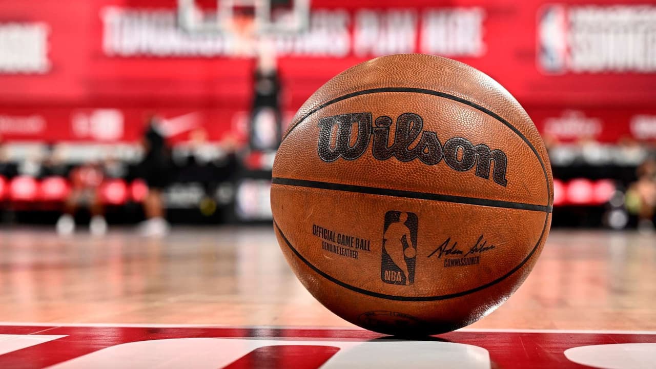 Primo piano di un pallone da basket ufficiale Wilson NBA che riposa su un campo in legno lucido con uno sfondo rosso.