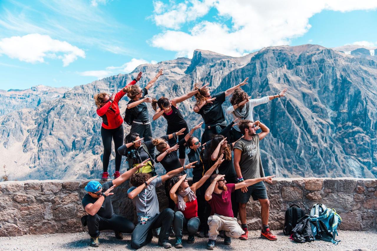 Un groupe WeRoad pose en formation 'dab' devant une chaîne de montagnes rocheuses sous un ciel bleu.