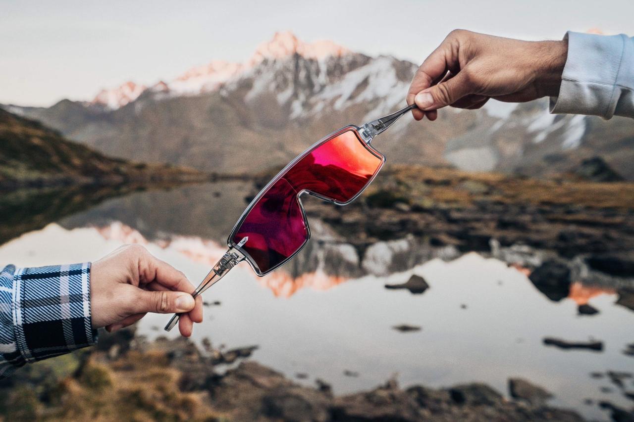 Eine rot getönte Sonnenbrille, gehalten von zwei Händen, mit Berglandschaft und See im Hintergrund.