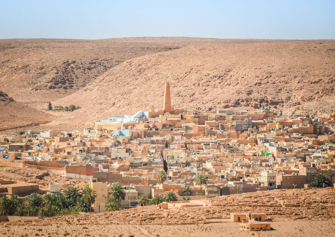 A desert village with sand-colored buildings and a central minaret is nestled at the base of a large, rocky hill under a clear sky.